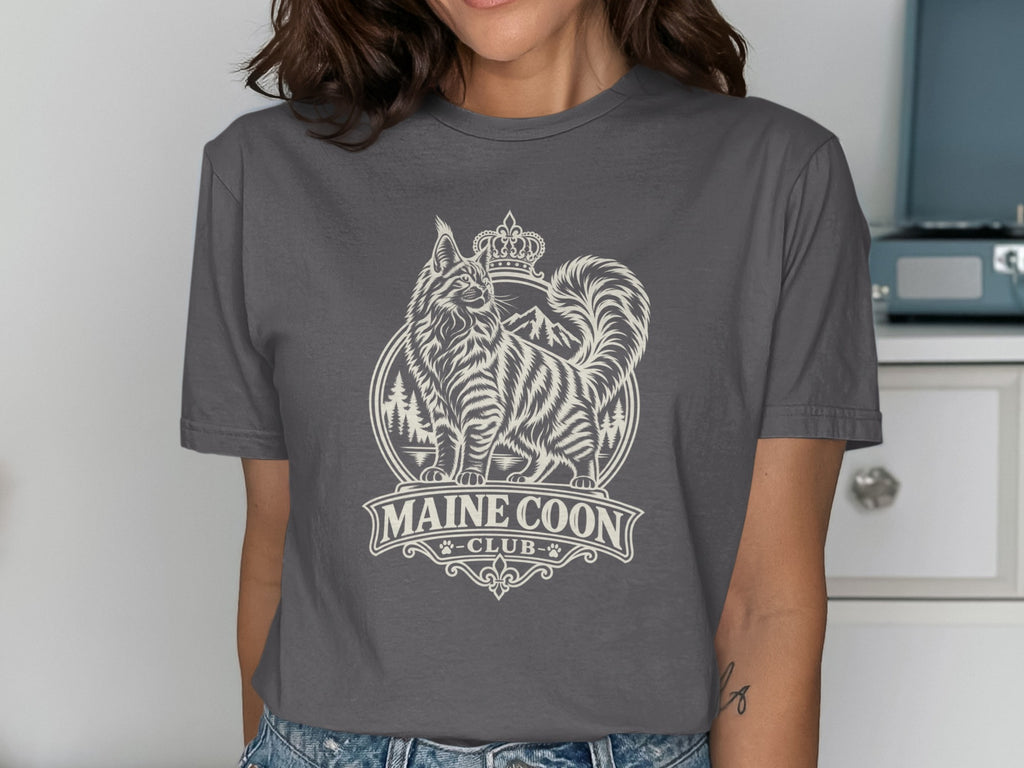 Maine Coon Club graphic with a crowned cat, mountains, and trees on a t-shirt.
