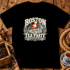 A black t-shirt features a vintage-style graphic centered on the chest, showing a crate of tea leaves with an orange traffic cone inside, surrounded by text that reads "Boston Tea Party, Nae Scotland, Nae Party."