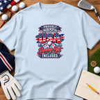 Light blue t-shirt featuring patriotic golf graphic and text surrounded by golf accessories on wood background.
